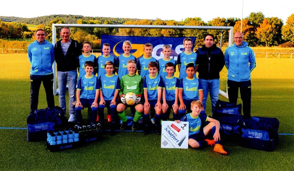 Eine Jugendfußballmannschaft posiert mit Trainern auf einem grünen Spielfeld vor einem blauen Banner.