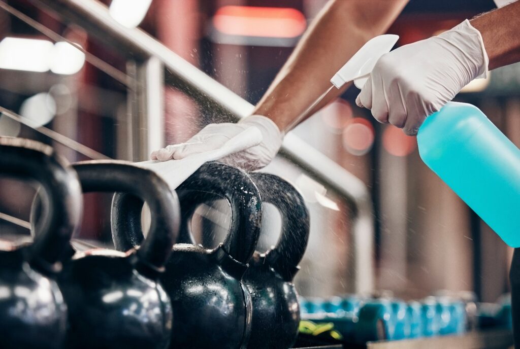Kettlebell, Reinigungsservice und Reinigung der Hände in einem Fitnessstudio mit Flüssigseife in einer Sprühflasche mit einem Tuch in Handschuhen. Hausmeister oder Reinigungskraft wischt schmutzige Bakterien von staubigen Gewichten oder Fitnessgeräten.