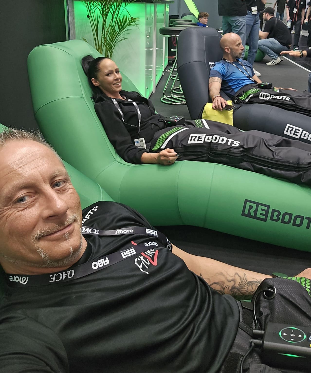 Drei Personen auf Relaxliegen bei einer Messe, lachen und entspannen, tragen Sportbekleidung in Schwarz.